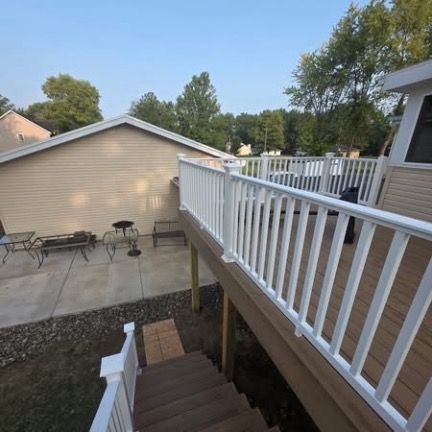 Elevated wooden deck with white railing, overlooking a concrete patio and a tan house exterior with a staircase below.
