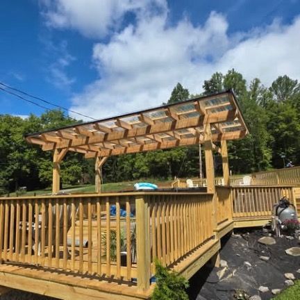A wooden deck featuring a pergola with a translucent, corrugated roof, set against a backdrop of trees and a blue sky.