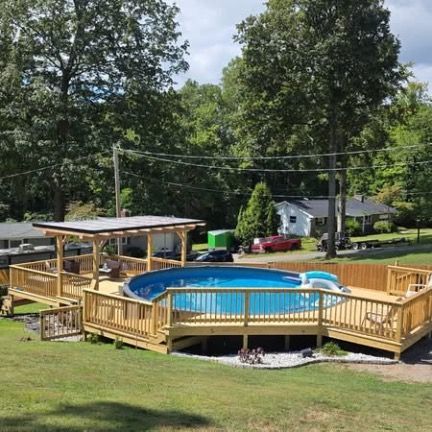 An above-ground swimming pool with a wraparound wooden deck and pergola, set in a grassy yard surrounded by trees.
