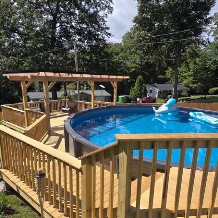 A round above-ground swimming pool with blue water and a pool float, surrounded by a wooden deck and a pergola.