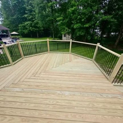 A wide, light-colored wooden deck with an angled layout and railings, overlooking a grassy backyard with trees.