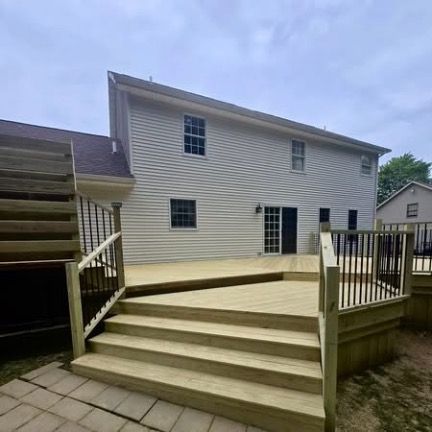 Newly constructed wooden deck with wide steps and black railings attached to the back of a light-sided suburban house.