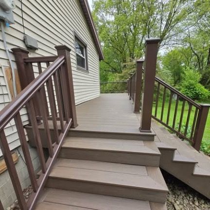 A brown composite deck with stairs and railing attached to the side of a house with beige siding, overlooking a yard.