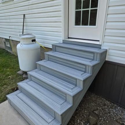 A set of five light grey composite stairs leading up to a white door on a house with light grey vinyl siding.