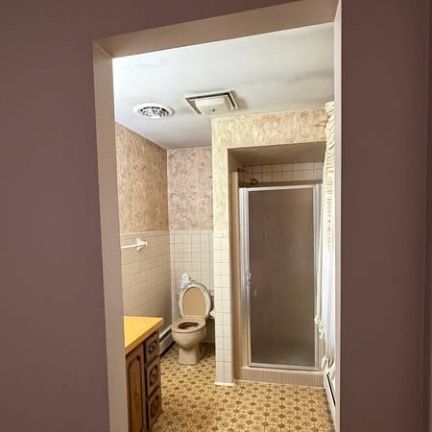 A view of a dated bathroom from a doorway, showing a beige toilet, shower stall, wood vanity, and patterned wallpaper.