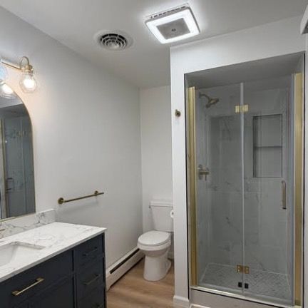 A bathroom with a dark blue vanity, marble countertop, gold hardware, toilet, and a glass-enclosed shower with gold trim.