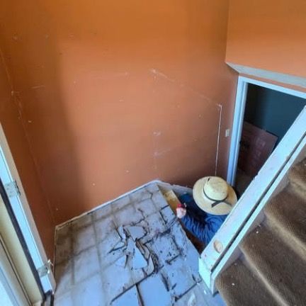 A person in a straw hat and blue shirt kneeling on a tiled floor near a staircase, working on home renovations.