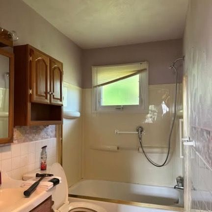 A bathroom features wood cabinets above a white sink, a toilet, and a bathtub with a handheld shower head under a window.