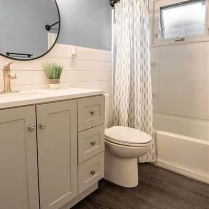 A small, modern bathroom with a light grey vanity, shiplap walls, round mirror, patterned shower curtain, and toilet.