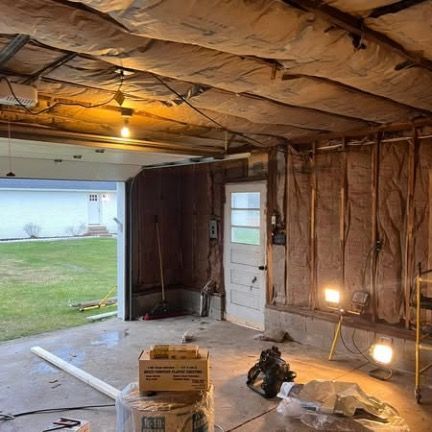 An unfinished garage with exposed fiberglass insulation on the walls and ceiling, a door to the outside, and work lights.