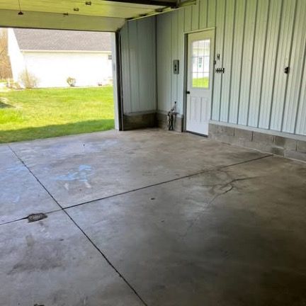 An empty, concrete-floored garage space with a white door and metal siding, looking out onto a grassy lawn.