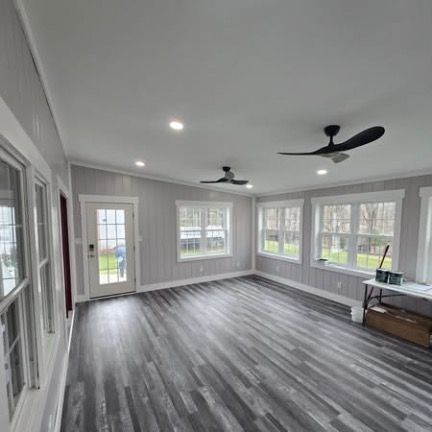 A sunroom with light gray vertical paneling, dark wood-look vinyl flooring, two black ceiling fans, and large windows.