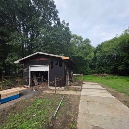 A detached garage under construction with black building wrap, scaffolding, and a concrete path in a wooded setting.