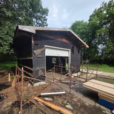 A detached garage under construction with black building paper, scaffolding, and stacked lumber in a grassy yard.