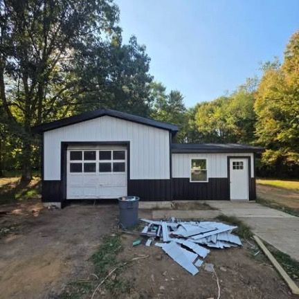 A white and black garage building with a roll-up door and a side entry door, set in a wooded area with debris on the ground.