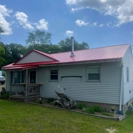 A single-story house with grey vinyl siding and a bright red metal roof, featuring a small wooden porch under a blue sky.
