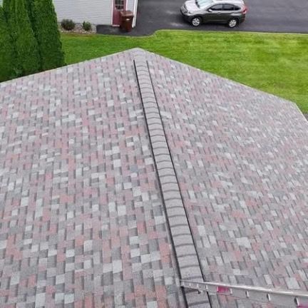A high-angle view looking down at a multi-colored shingled roof with a central ridge vent, and a ladder resting on the edge.