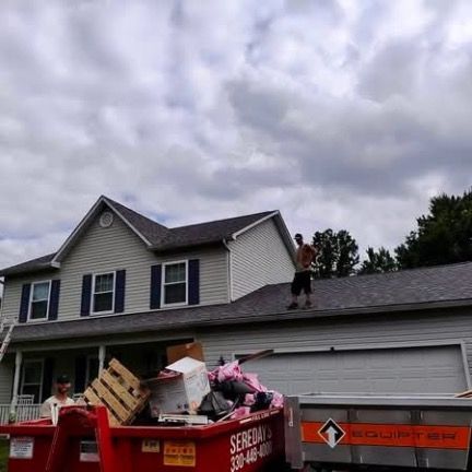 A worker stands on the roof of a house during a renovation project, with a large dumpster filled with debris below.