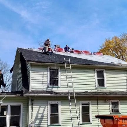 Four people work on a house roof on a sunny day with new roofing materials staged near the peak.