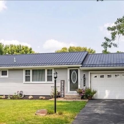 A single-story gray house with a dark metal roof, a white garage door, and a front lawn with a black lamp post.