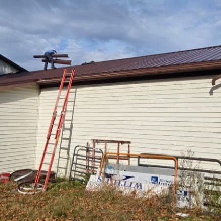A worker stands on a brown metal roof carrying materials next to an orange ladder leaned against a beige building.