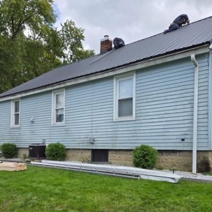 Two workers on a light blue house roof, installing a dark metal roof, with green shrubs and grass in the foreground.