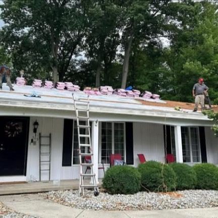 Workers install shingles on a white house roof, with an extension ladder leaning against the porch.