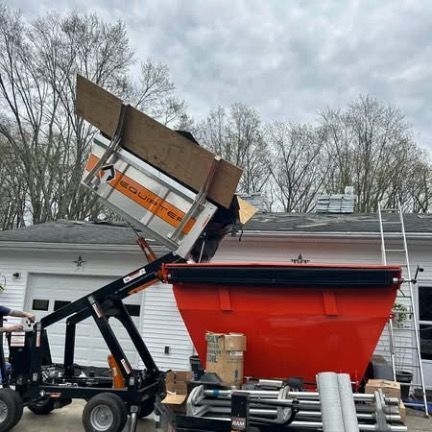 A dump trailer is tilted, unloading building materials into a large red dumpster in front of a house.