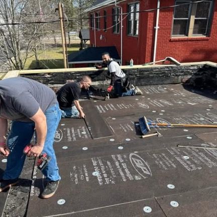 Three construction workers install roofing material on a flat roof next to a red brick building.