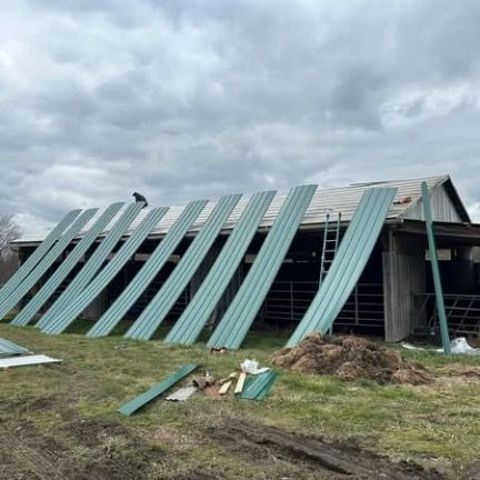 A person stands on a barn roof as green corrugated metal panels are being installed over existing roofing materials.