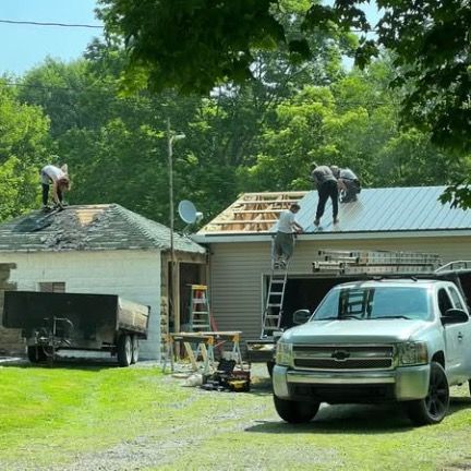 Workers install a metal roof on a garage next to a smaller building with a trailer parked on a grassy lot.