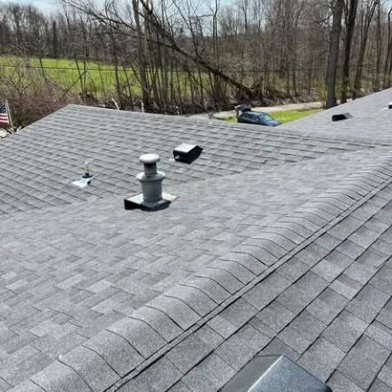 A high-angle view of a gray shingled roof with plumbing vents and a dark car visible in the background trees.