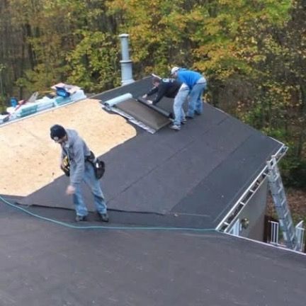 Three workers in casual clothes install black roofing underlayment onto the wooden deck of a residential roof.