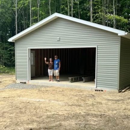 Two people stand in the open doorway of a new, gray-sided garage under construction in a wooded area.