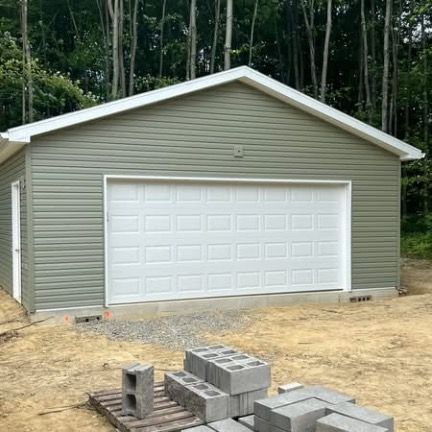 A detached gray garage with a white door and trim, situated on a dirt lot with stacked concrete blocks in the foreground.