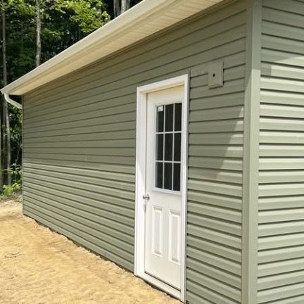 Side view of a sage green shed with white trim and a single white door featuring a small window, set against a tree line.