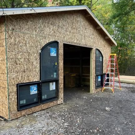 An unfinished wooden shed under construction featuring arched black-framed windows and a ladder leaning against the wall.