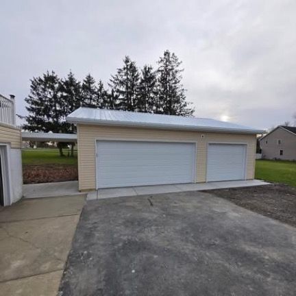 A beige detached garage with two white doors and a metal roof, connected to a house by a covered walkway.