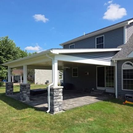 A gray house with a stone-columned patio cover extending over a stone paver patio on a grassy lawn under a blue sky.