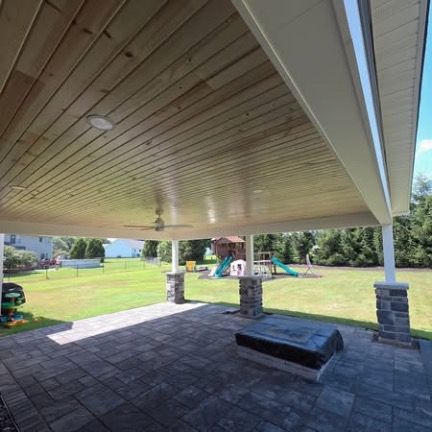 A covered stone patio with a wood-plank ceiling overlooks a grassy backyard with a playset and a blue slide.