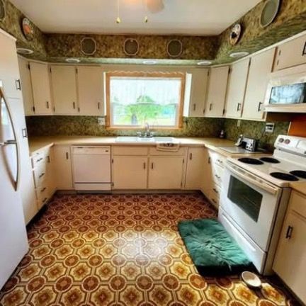 A dated U-shaped kitchen with white cabinets, patterned wallpaper, a green dog bed on the floor, and a vinyl tile floor.