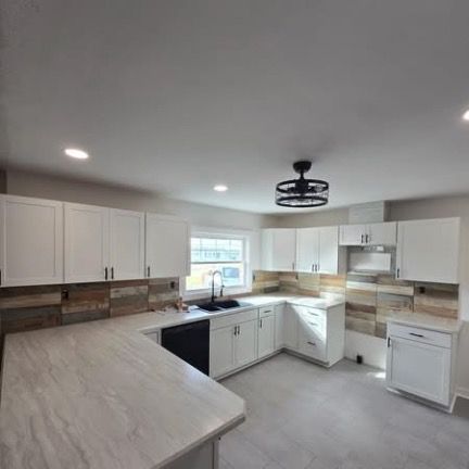 A bright kitchen featuring white cabinets, wood-look backsplash, a light-colored countertop, and a black ceiling fan.