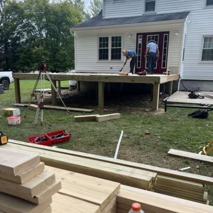 Two people work on a wood deck frame under construction outside a two-story white house with a dark red door.