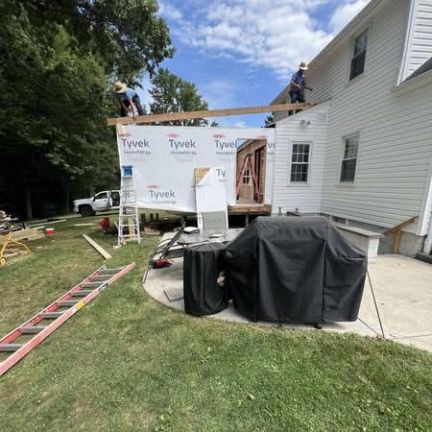 Two people work on a house addition under construction, featuring white Tyvek siding, a wooden frame, and a patio area.