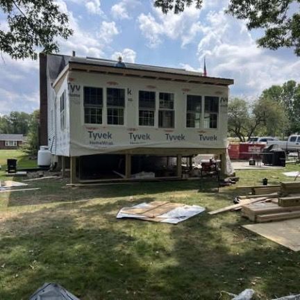 A house addition under construction with Tyvek wrap and new windows, viewed from a grassy backyard on a sunny day.