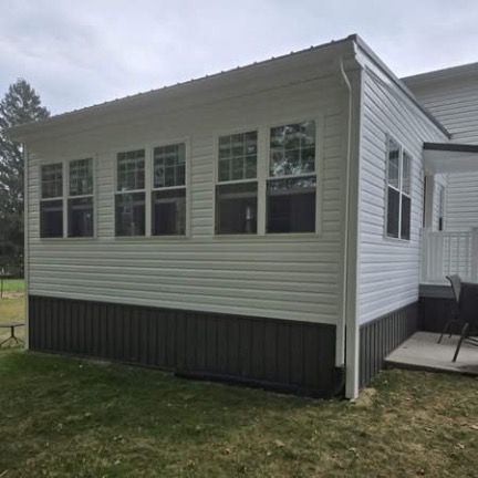 A white sunroom addition with a dark gray metal skirting, multiple windows, and a metal roof attached to a house.