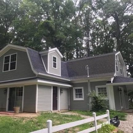 A two-story, gray, gambrel-roofed house with white trim, a dormer window, and a white fence in a wooded setting.