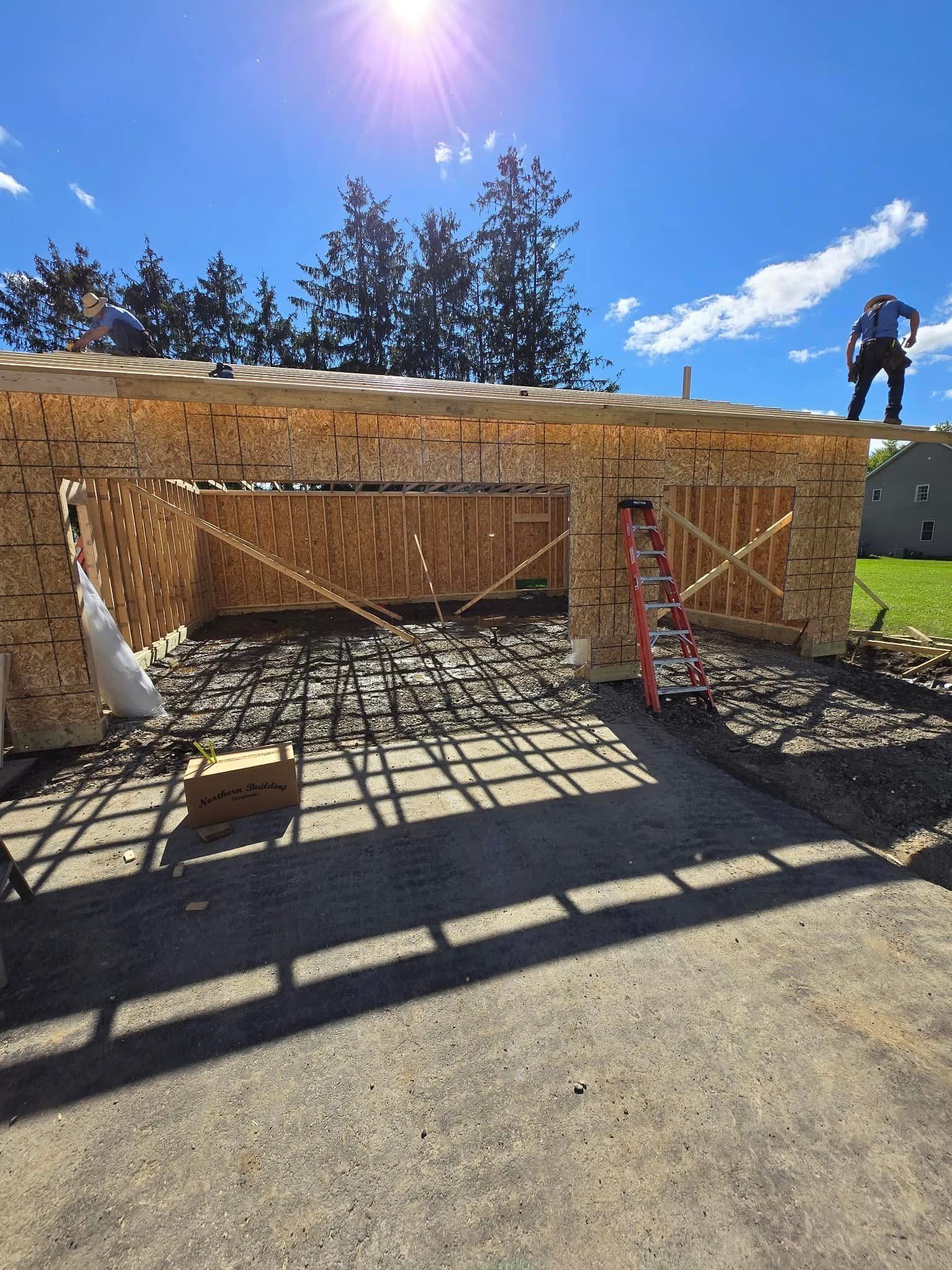 A construction site showing a wooden garage frame under a sunny sky with two workers on the roof.