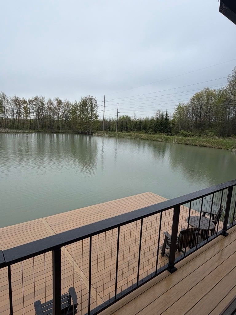 Pond view from a patio with black railing, tan deck, and trees lining the far shore under an overcast sky