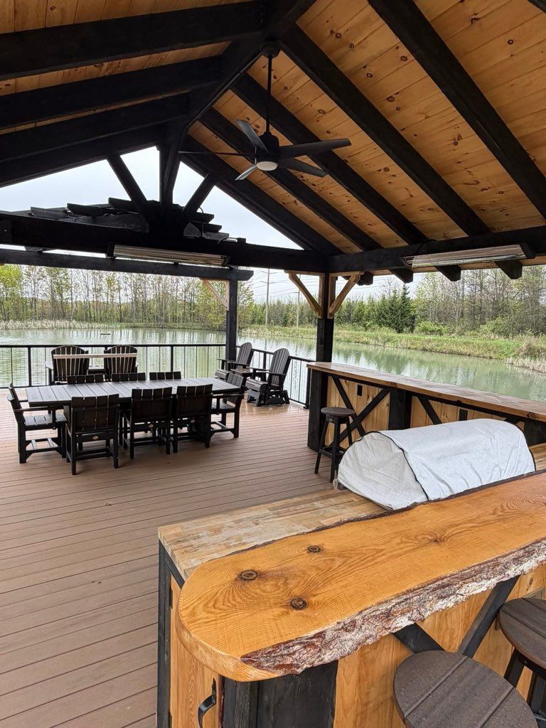 Covered outdoor deck with picnic table, benches, and a wooden roof overlooking a lake and trees